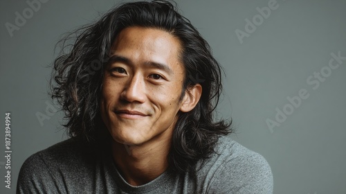 Smiling Portrait: A man with long, dark hair and a casual gray shirt poses against a simple gray background, radiating confidence and warmth with his engaging, happy expression.