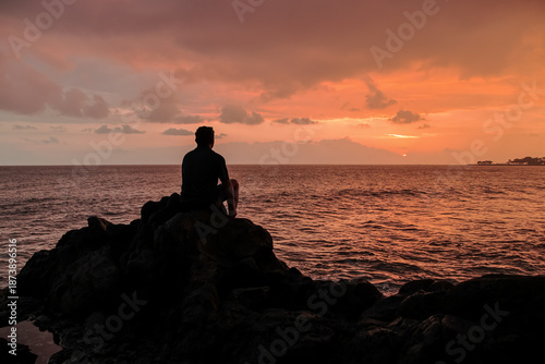 Man sitting on rocks gazing at ocean during sunset with vivid colors for travel and relaxation concepts using natural light