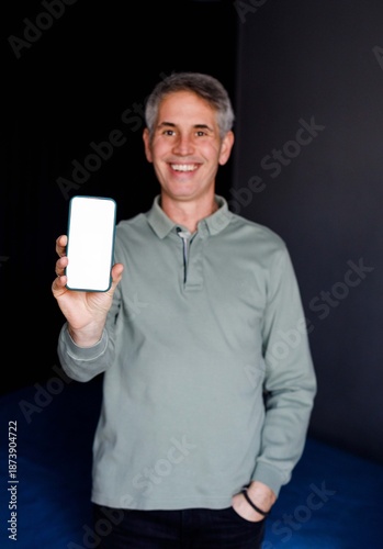 Smiling man holding smartphone with blank screen while working on laptop at home office.