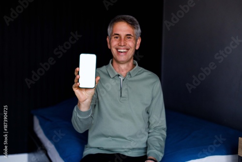 Smiling man holding smartphone with blank screen while working on laptop at home office.