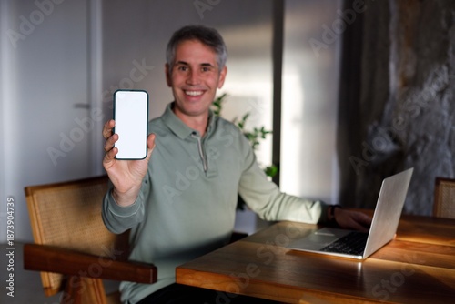 Smiling man holding smartphone with blank screen while working on laptop at home office.
