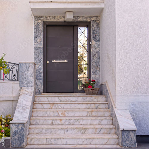 Minimalist home entrance featuring  dark grey security door framed by marble. Elegant white marble stairs lead to a sleek entryway accented with a charming basket of red flowers in Athens, Greece.