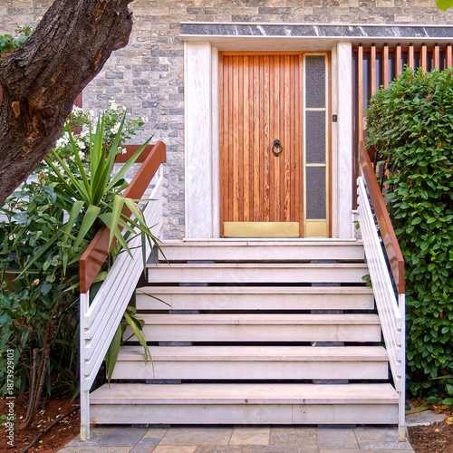 Elegant home entrance featuring white marble stairs leading to a vertical slat wooden door. A sophisticated blend of natural textures, stone masonry, and lush Mediterranean greenery in Athens, Greece.