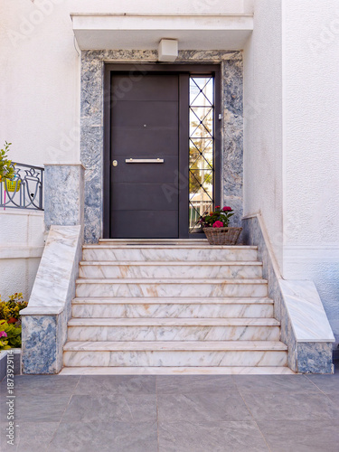 Elegant home entrance in Athens featuring a dark grey security door framed by blue-veined marble. White marble stairs lead to a modern entryway accented with red flowers.