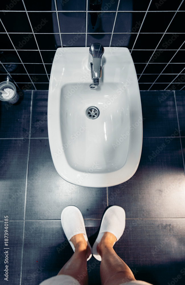 Fototapeta premium A clean bidet in a modern bathroom. White ceramic fixture on dark tiled floor. A pair of white slippers are visible in the foreground.