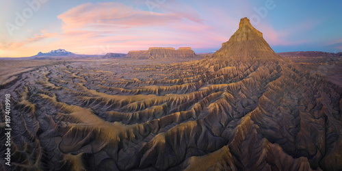 Aerial view of stark, eroded badlands bathed in the warm glow of sunset, with Factory Butte rising majestically against the pastel sky, Hanksville, Utah, United States.