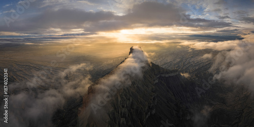 Aerial view of a rugged, dark mountain peak piercing through a sea of ethereal clouds, bathed in the golden light of the setting sun, Hanksville, Utah, United States.