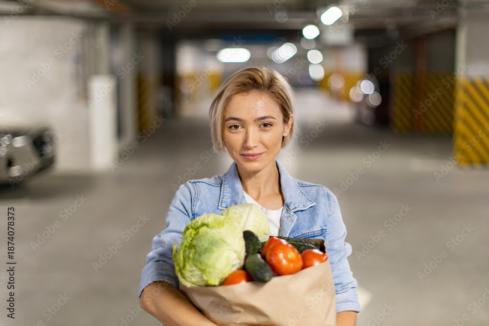 Fototapeta premium A woman smiles while holding a bag of fresh produce in a parking garage, ready to head home