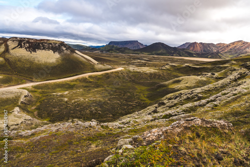 View of a winding road cutting through a dramatic landscape of mossy hills under a cloudy sky, creating a sense of vastness and adventure, Landmannalaugar, The Central Highland, Iceland.