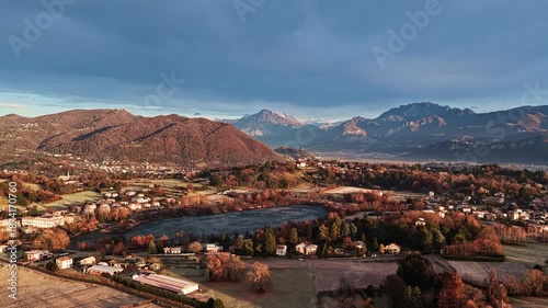 Aerial drone shot with a smooth circular movement to the left, showing a frozen lake fully surrounded by winter fields, villages and mountains in northern Italy at sunrise.