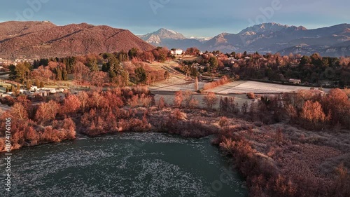 Aerial drone shot rising from low to higher altitude over a frozen lake, revealing winter countryside and mountains in soft sunrise light.