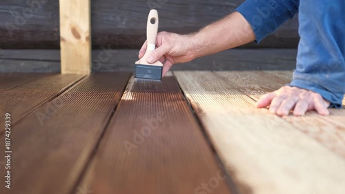 Brown oil for protection of wood by staining. Painting a larch for protecting and pests. Woodworker applying finish to terrace deck board. Repairman varnishing with brush wooden plank floor surface.