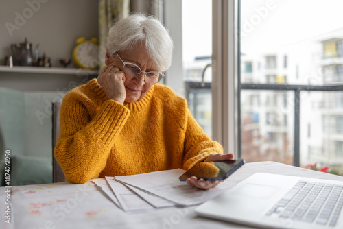 Senior woman holding mobile phone while reading paper documents at home
