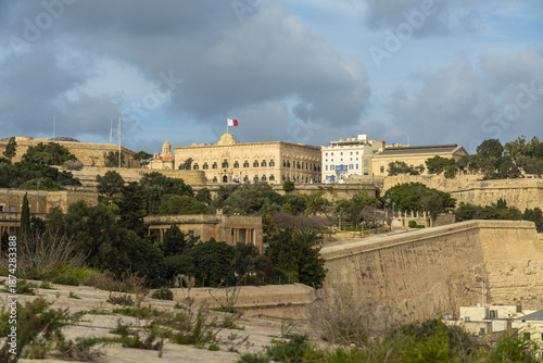 View of imposing stone fortifications crowned by governmental buildings with the Maltese flag fluttering against a cloudy sky, Valletta, Malta.