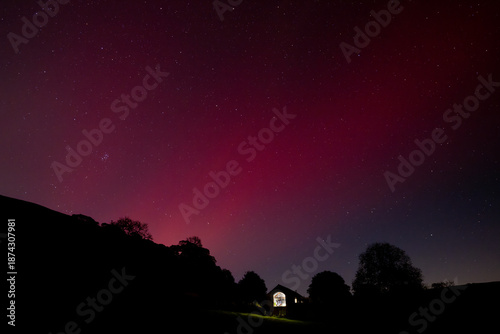 The aurora borealis glows in the night sky above a silhouetted house and trees, creating a magical and serene scene.