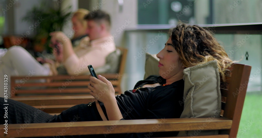 Fototapeta premium Young woman relaxing on wooden bench using smartphone indoors, with two people in background using mobile devices in a casual shared workspace