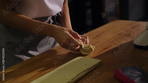 Baker shapes rolled dough into Swedish cinnamon buns, twisting and folding each piece by hand on a work surface. Clear view of the forming process, fresh dough texture, warm bakery setting.