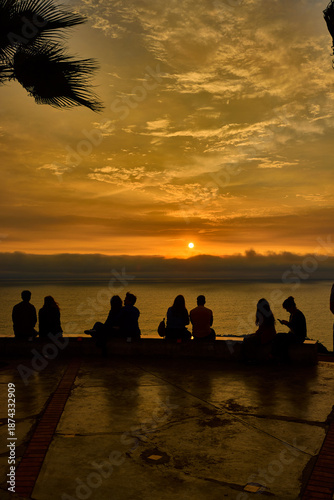 As the sun begins its descent over the Pacific Ocean, locals and travelers alike gather along the cliffs of Miraflores, Lima. Framed by towering palms, the sky transforms into a canvas of amber 