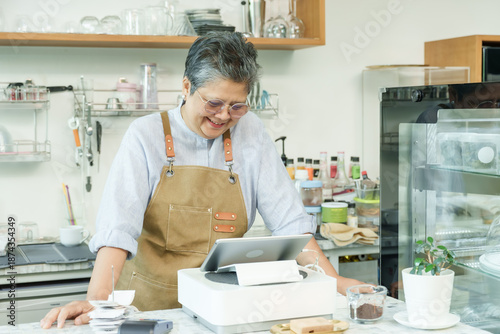 Billede på lærred Asian senior female cafe owner smiling leaning on counter using tablet enjoying working confidently running independent small coffee shop demonstrating post-retirement happiness in business