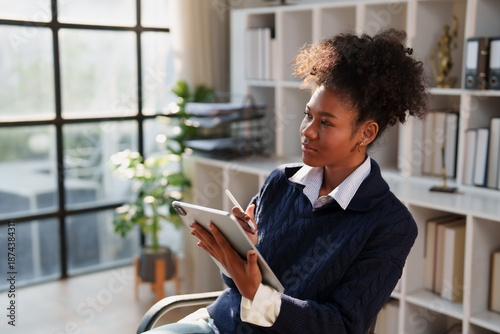 Professional young businesswoman sitting in a modern office, concentrating and looking away while using a digital tablet with a stylus pen, working on new ideas