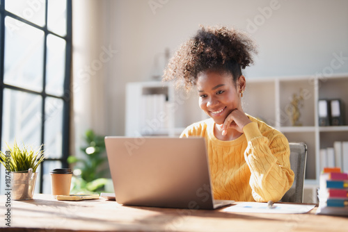 Young happy African American woman smiling while looking at a laptop screen, engaging in online work from a modern office, enjoying remote productivity