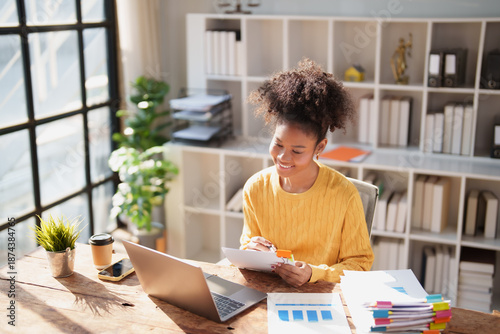 Tableau sur toile Young Black woman at desk happily reviewing paperwork and financial charts, orga