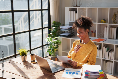 Young happy African American woman reviewing financial documents and planning strategy at her desk, with a laptop and organized files in a modern office environment