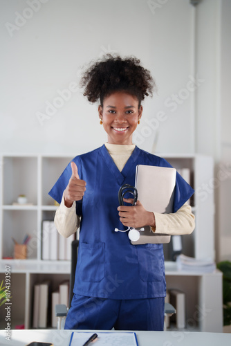 Young African American woman wearing blue scrubs, smiling confidently and giving a thumbs up gesture while holding a laptop and stethoscope in a healthcare setting