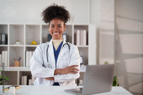 African American woman doctor wearing a white lab coat and stethoscope, smiling confidently while standing with crossed arms at her desk in a modern healthcare clinic