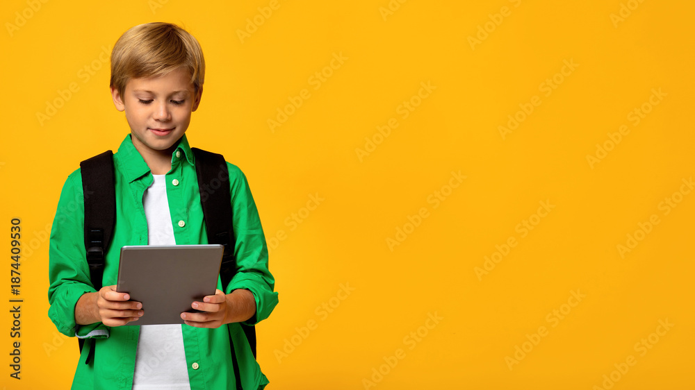 Obraz premium Smiling caucasian teen boy student with backpack typing on tablet and watching educational video, isolated on yellow background, copy space. Online education, remote schooling, distance learning class