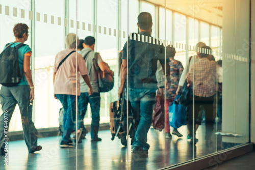 Photograph through large clear glass wall with blurred image of departing passengers and luggage walking airplane boarding corridor from terminal to plane in background. Selective focus on glass.