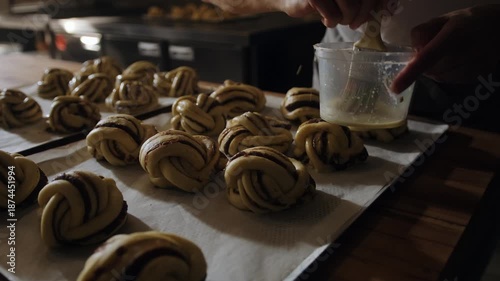 Close-up of an artisan baker brushing egg wash onto Scandinavian cinnamon and cardamom morning buns before baking. Traditional Nordic pastry process, morning pastry, natural light, hygge mood. Small  