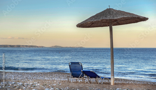 straw umbrella and sun loungers on a rocky beach at sunset in front of blue s...