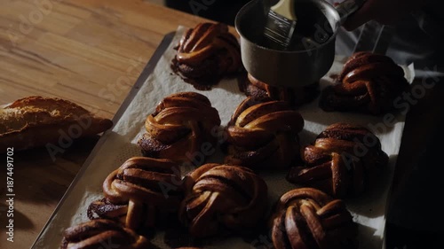 Baker glazing freshly baked cinnamon buns with sweet syrup. Golden crust shines as the syrup slowly absorbs into the warm pastry, artisan quality handcrafted bakery. Hygge atmosphere, premium bakery.