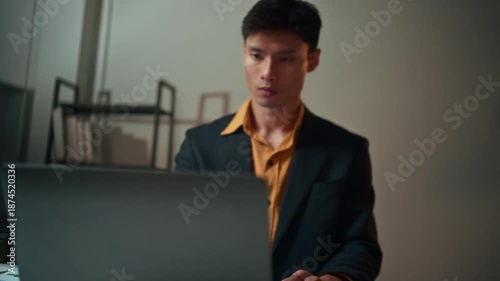 Focused asian businessman using a laptop and calculator while working late. Overworked employee analyzing financial data and charts at his desk