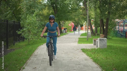 Wallpaper Mural woman cycling toward camera on park path, smiling with helmet and confident posture, bright daylight, playful energy, balanced pedaling, approaching walkway with trees and benches, cheerful commuting Torontodigital.ca