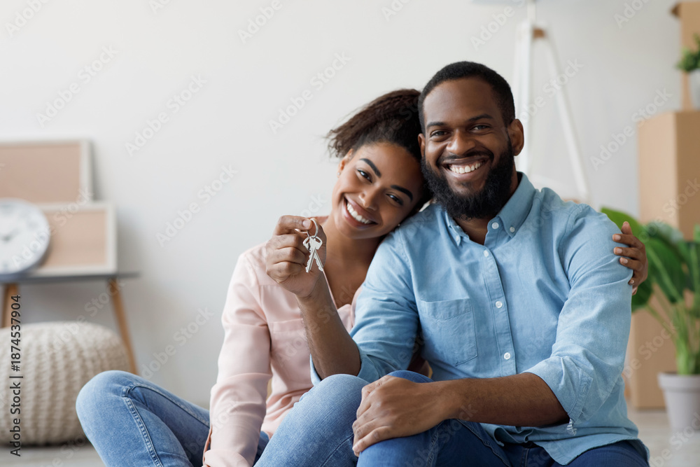 Obraz premium Happy young black couple hugging and showing keys to new apartment with cardboard boxes, looking at camera after relocation. Buying own real estate, renting apartment, moving, positive emotions