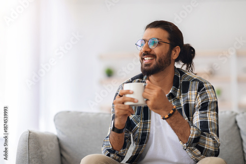 Billede på lærred Cheerful young indian man with coffee mug looking at copy space, sitting on couc