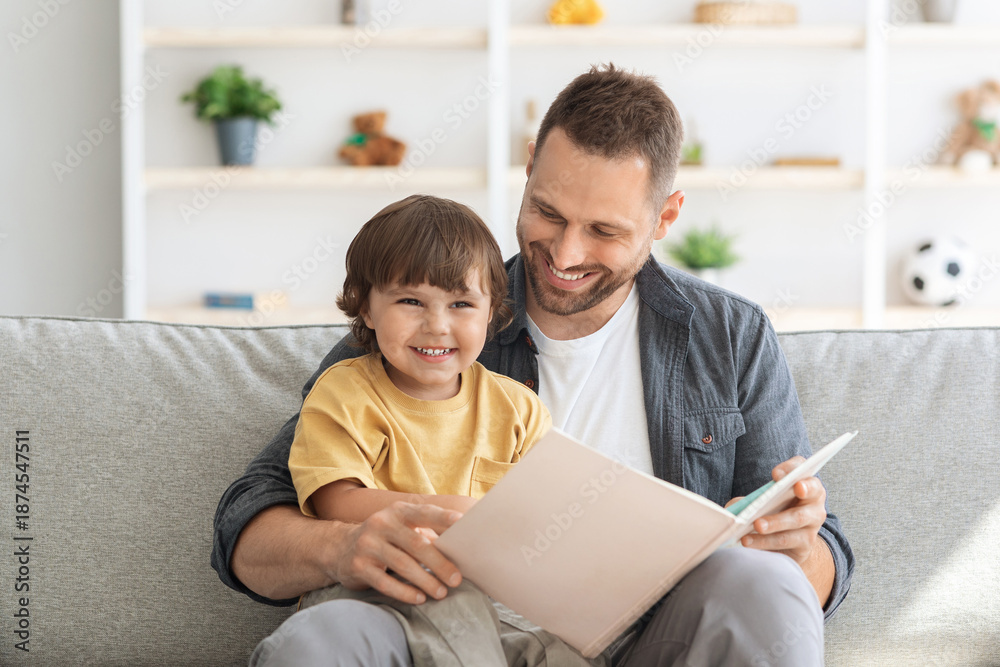 Fototapeta premium Family and upbringing concept. Close up portrait of happy little boy enjoying storytelling time with his daddy, sitting in armchair with big book
