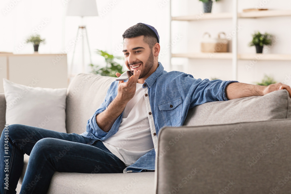 Fototapeta premium Phone Call, Mobile Communication And Voice Assistant Concept. Smiling Bearded Man In Kipa Talking On Smartphone Using Loudspeaker Mode, Guy Sitting On Comgortable Couch, Selective Focus