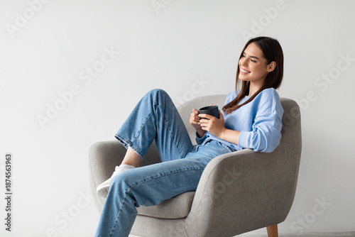 Canvas Print Portrait of happy young woman sitting in armchair with cup of hot aromatic coffee, relaxing on white studio background