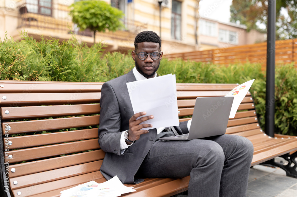 Fototapeta premium Business career concept. Serious african american entrepreneur working on laptop and reading documents, sitting on bench near office building. Black businessman holding papers