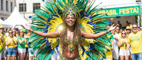 a Black woman with braided hair, wearing a large, colorful Brazilian carnival headdress, green and yellow feathers, and a matching costume, with arms outstretched, smiling.