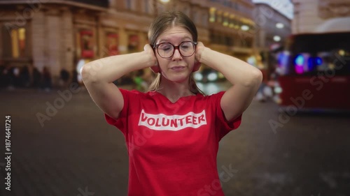 Blonde volunteer woman covers ears with hands on busy city street at night amid neon lights; stress noise overload tension.