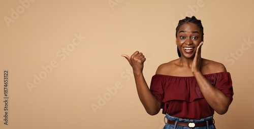 Surprised afro-american millennial woman in casual pointing at copy space for advertisement and touching her face, showing amazing deal or shocking news, beige studio background, panorama
