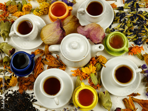 Porcelain cups and a teapot on a white table among various dried plants. Ingredients for making herbal and flower tea, top view.
