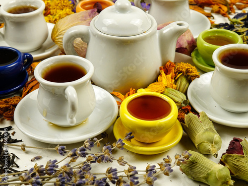 Porcelain cups and a teapot on a white table among various dried plants. Ingredients for making herbal and flower tea, side view.