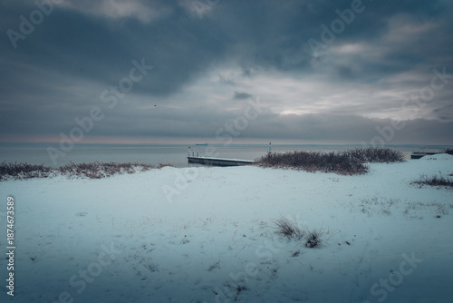 A scenic winter view from Raa Vallar in Helsingborg, Sweden. The image shows a snow-covered coastline and beach leading toward a calm sea with a wooden pier.
