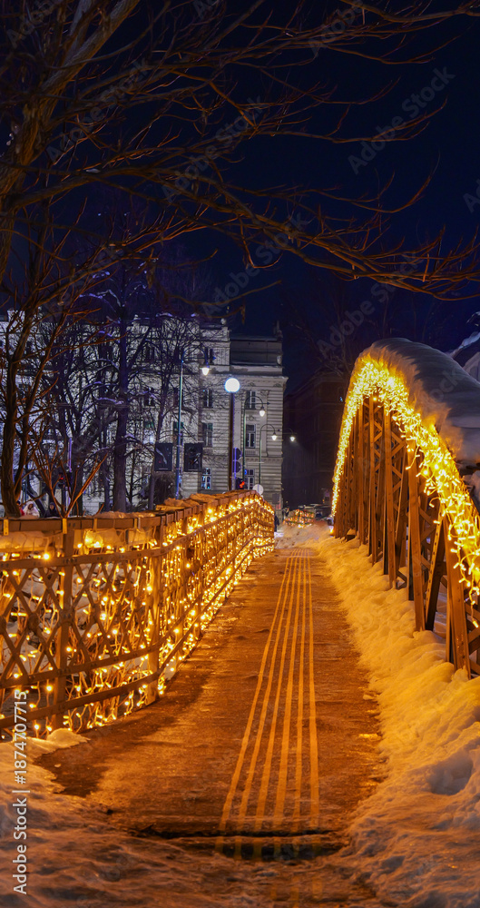 Fototapeta premium Pedestrian bridge decorated with festive lights during a snowy winter night, creating a cozy and atmospheric urban walkway.