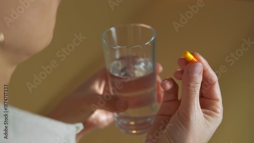 Woman taking medication pill with a glass of water. Unrecognizable woman taking a daily dose of medication, swallowing an orange and yellow capsule with a full glass of water for health, wellness